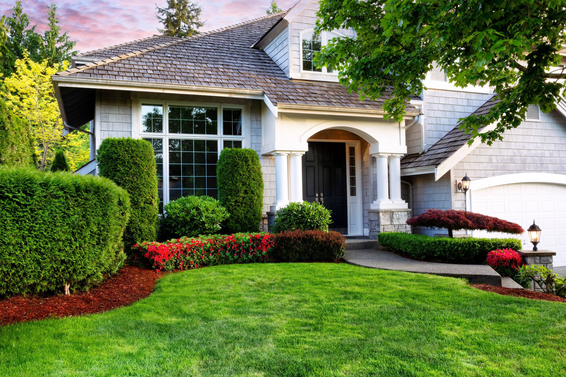 Elegant two-story home with white columns, black front door, manicured lawn, red flower beds, and green hedges