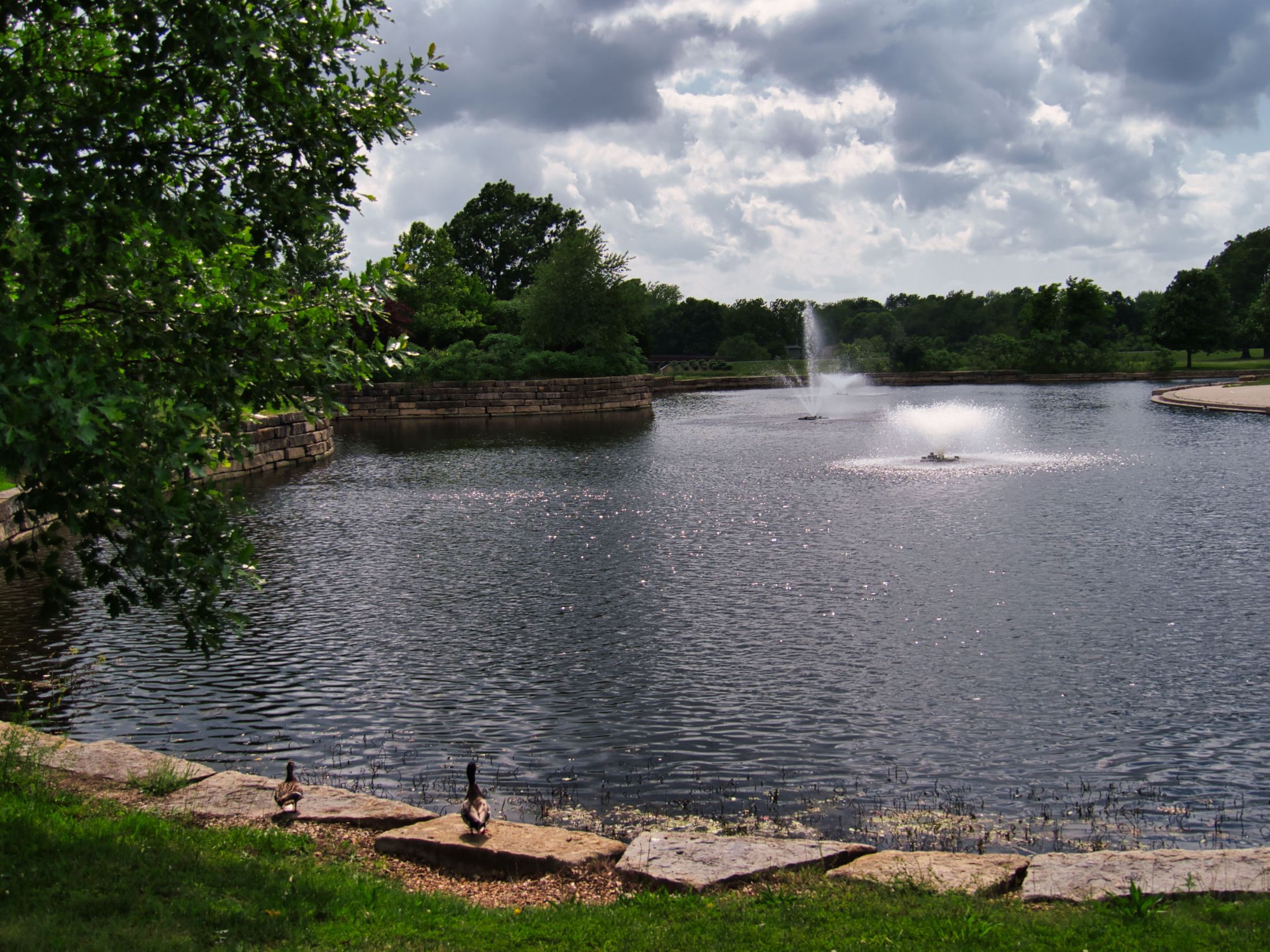 Scenic lakeside view with fountain, brick walls, lush trees, geese on sandy shore, and cloudy sky