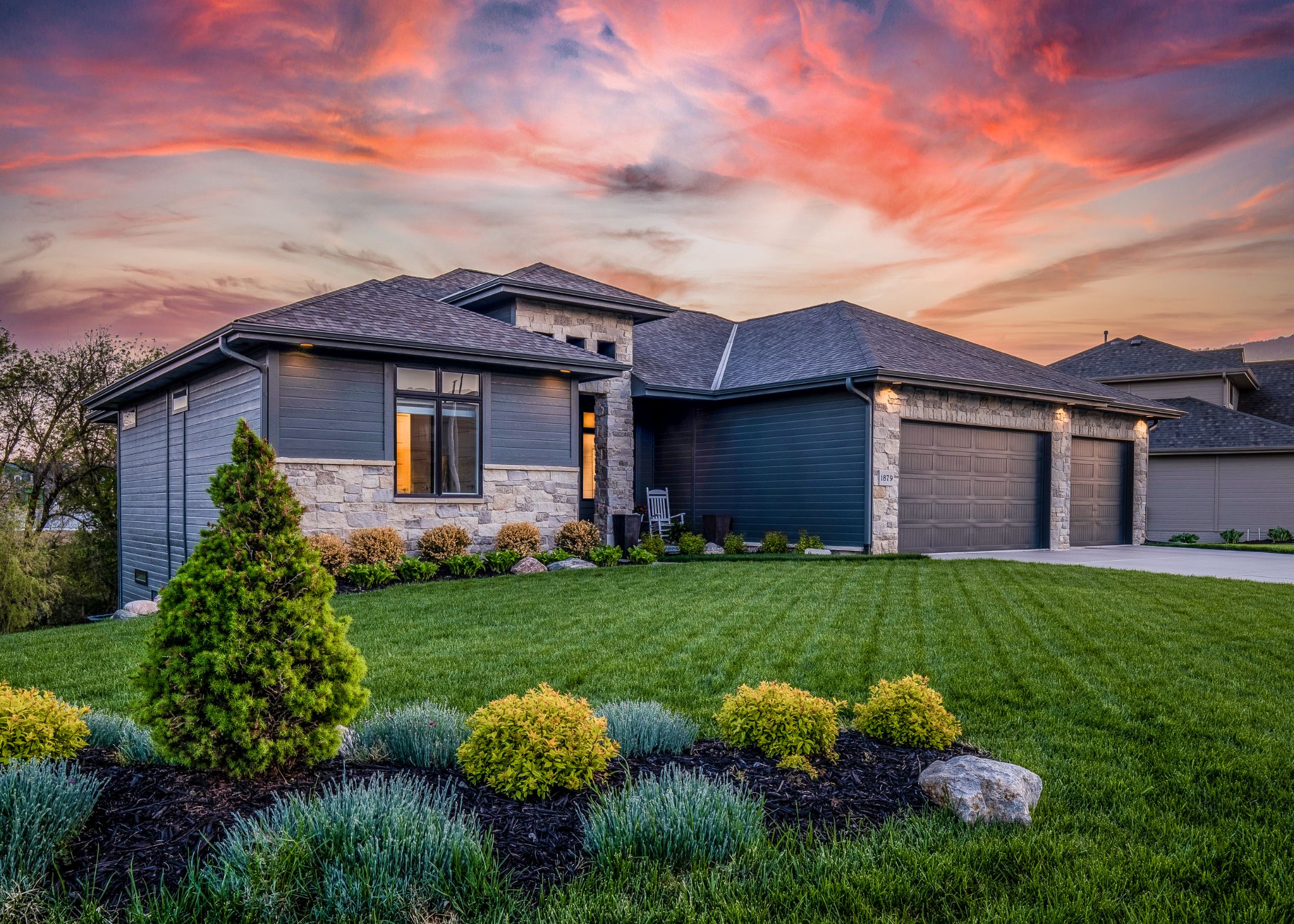 Modern home with stone and blue siding, three-car garage, manicured lawn and landscaping at sunset
